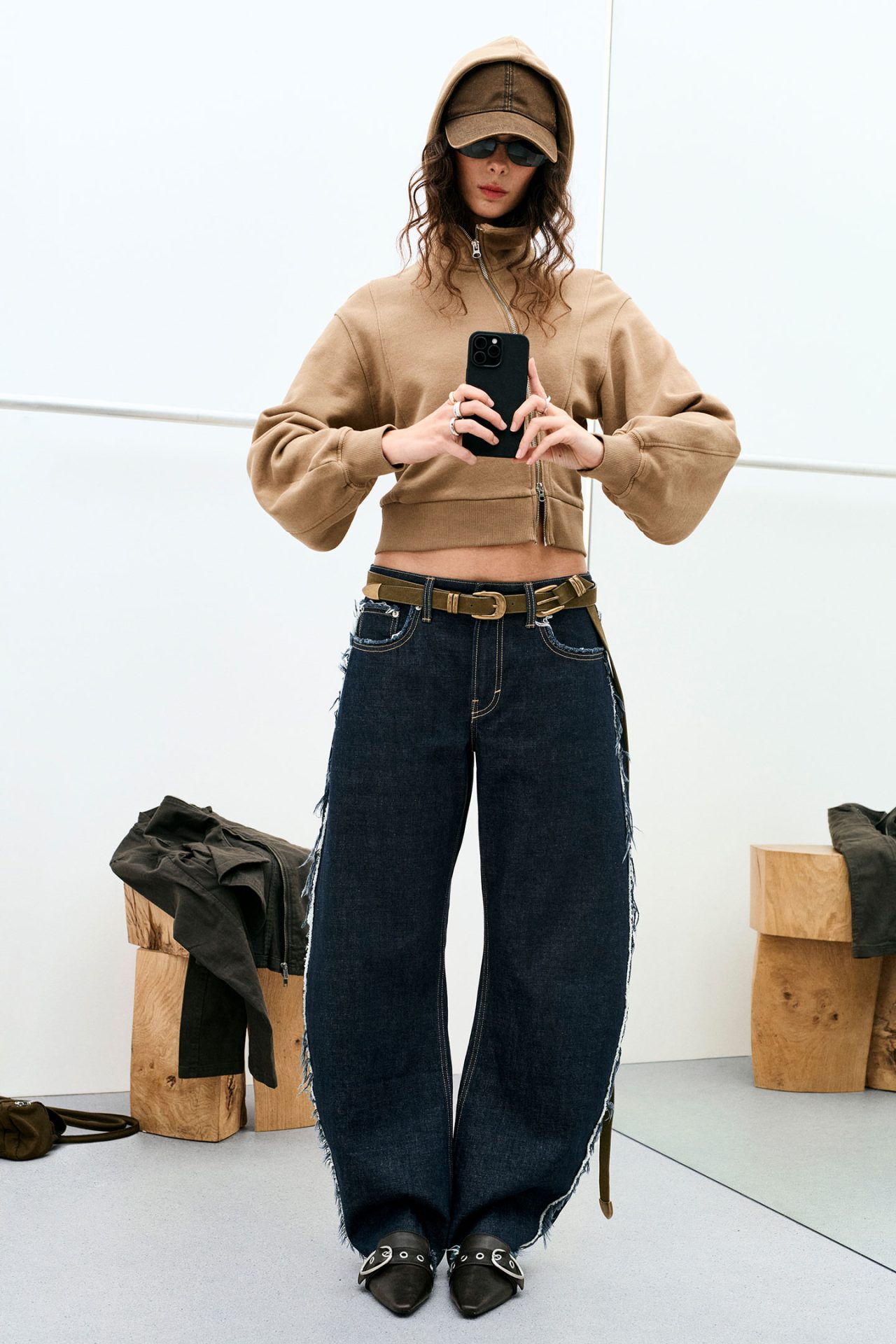 Model wearing a cropped brown zip-up hoodie and oversized dark wide-leg jeans with raw edge detailing, styled with a cap, sunglasses and pointed shoes, showcasing Weekday’s directional silhouettes.
