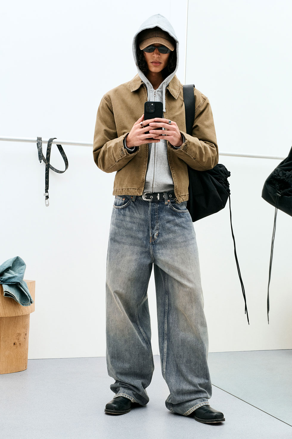 Model in a layered grey hoodie and brown jacket paired with loose light-wash wide-leg jeans, accessorised with sunglasses and a shoulder bag, reflecting Weekday’s relaxed, contemporary street style.