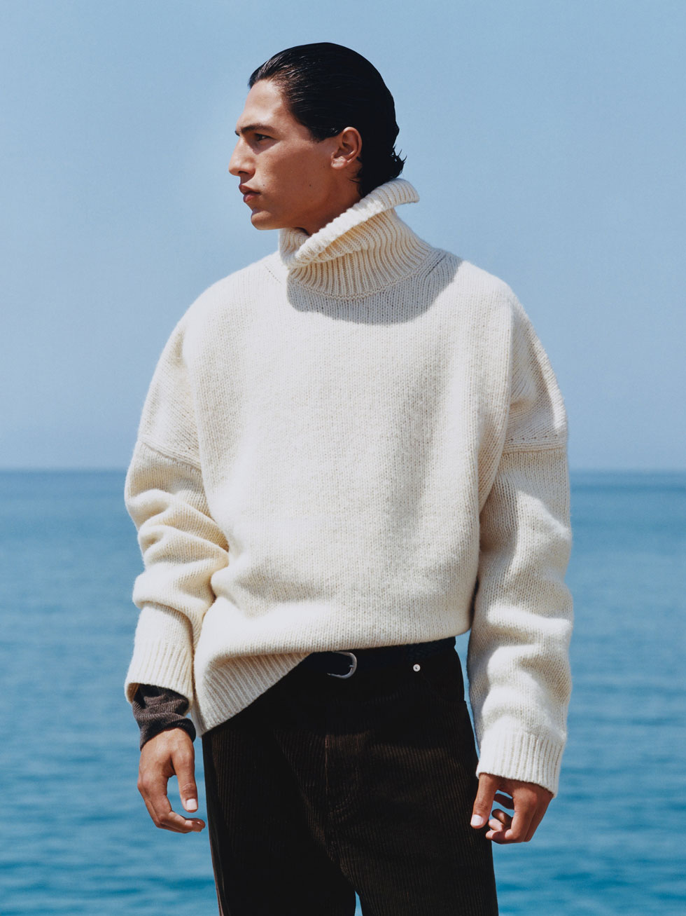 Model in an oversized cream turtleneck sweater and dark trousers stands by the ocean, looking sideways