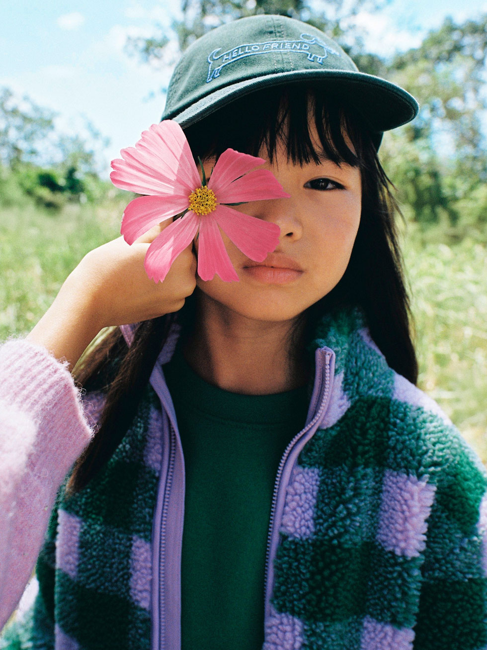 Model in a green baseball cap and purple-green check fleece holds a pink flower over one eye in a sunny field.