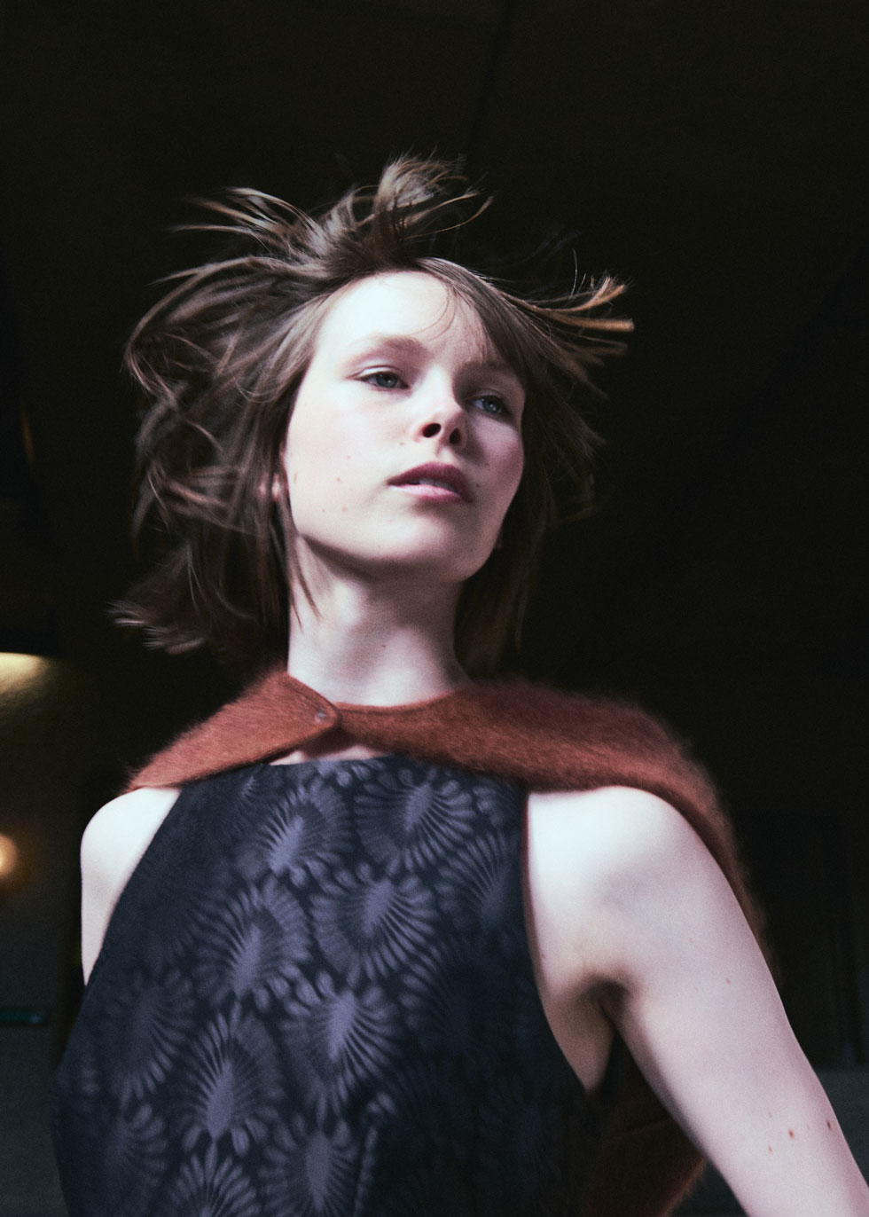 Close-up with motion: model’s hair tousled, wearing a black patterned dress with a rust fuzzy capelet against a dark backdrop.