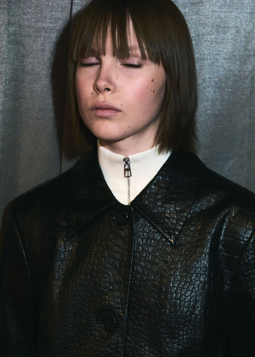 Portrait: model in a croc-embossed black coat over a white zip-neck top, eyes closed.