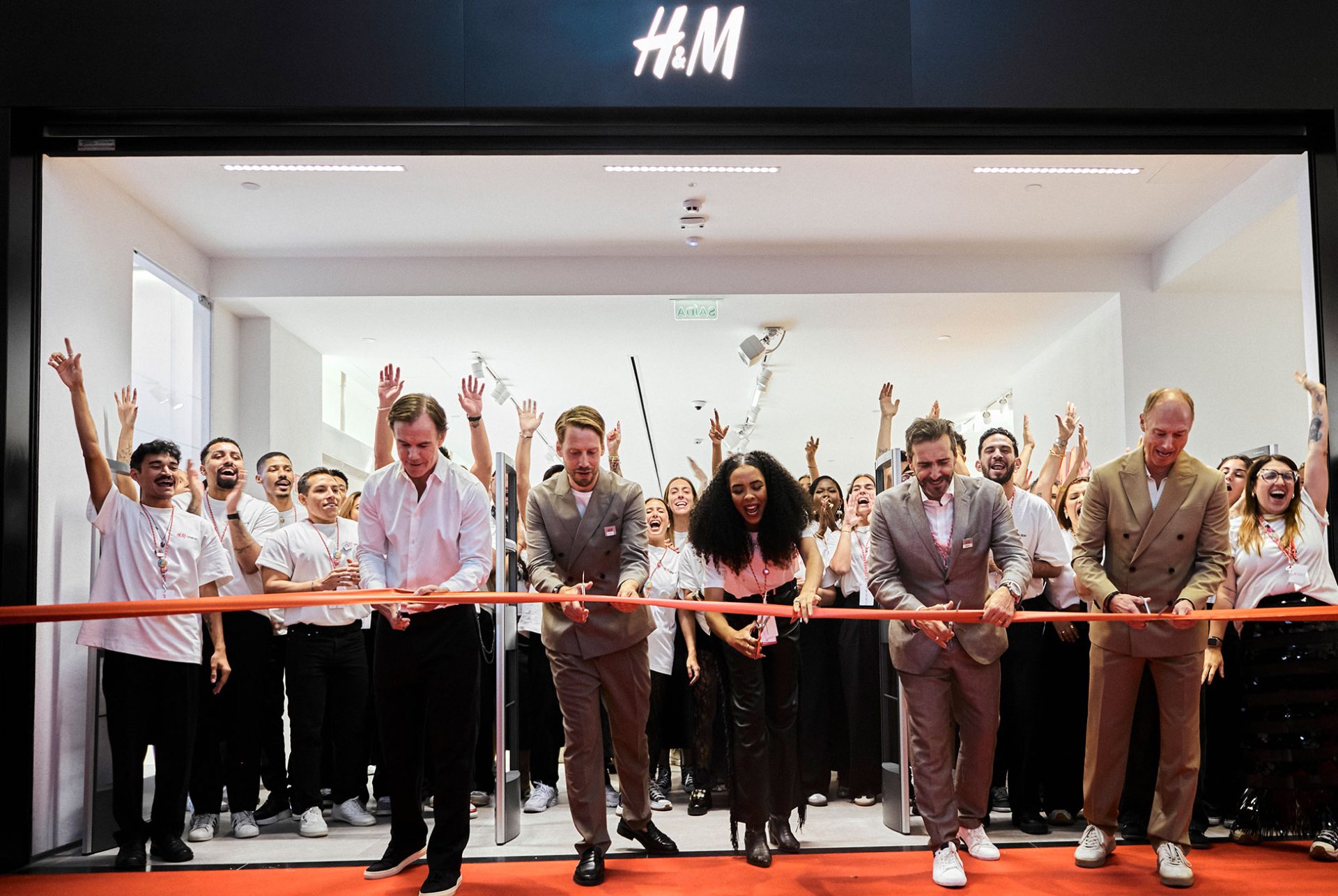 Group of people celebrating the opening of the first H&M store in Brazil, with a ribbon-cutting ceremony in front of the brightly lit entrance and the H&M logo displayed above.
